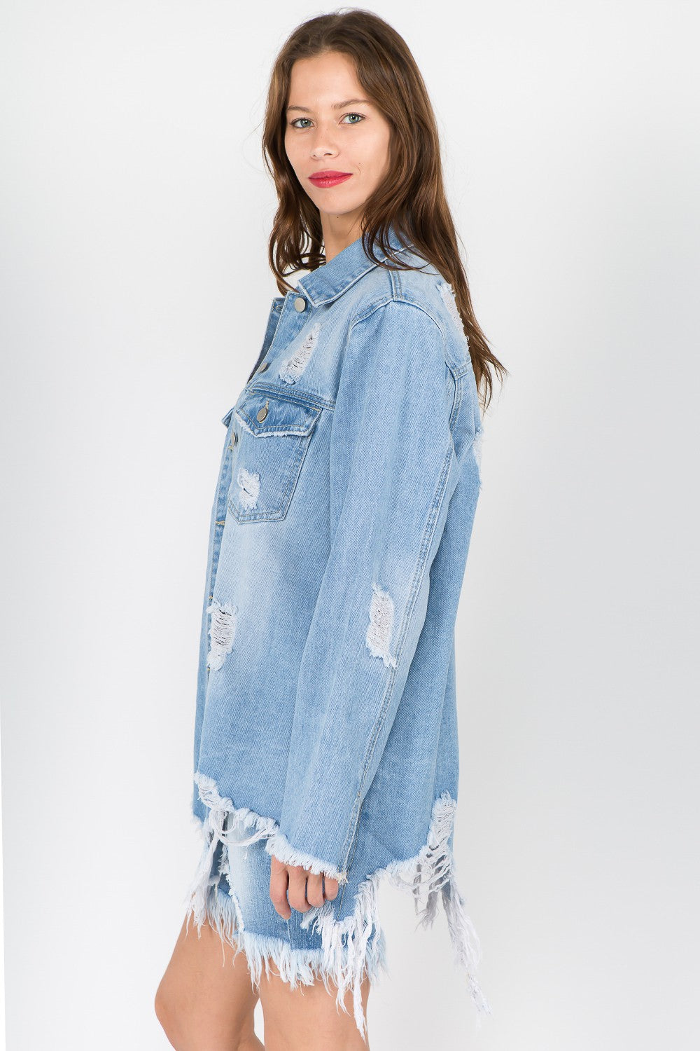 A woman sporting an American Bazi Distressed Frayed Hem Denim Jacket in light blue, paired with a white top and ripped denim shorts, stands against a plain background, exuding a vintage vibe with the frayed hem details.