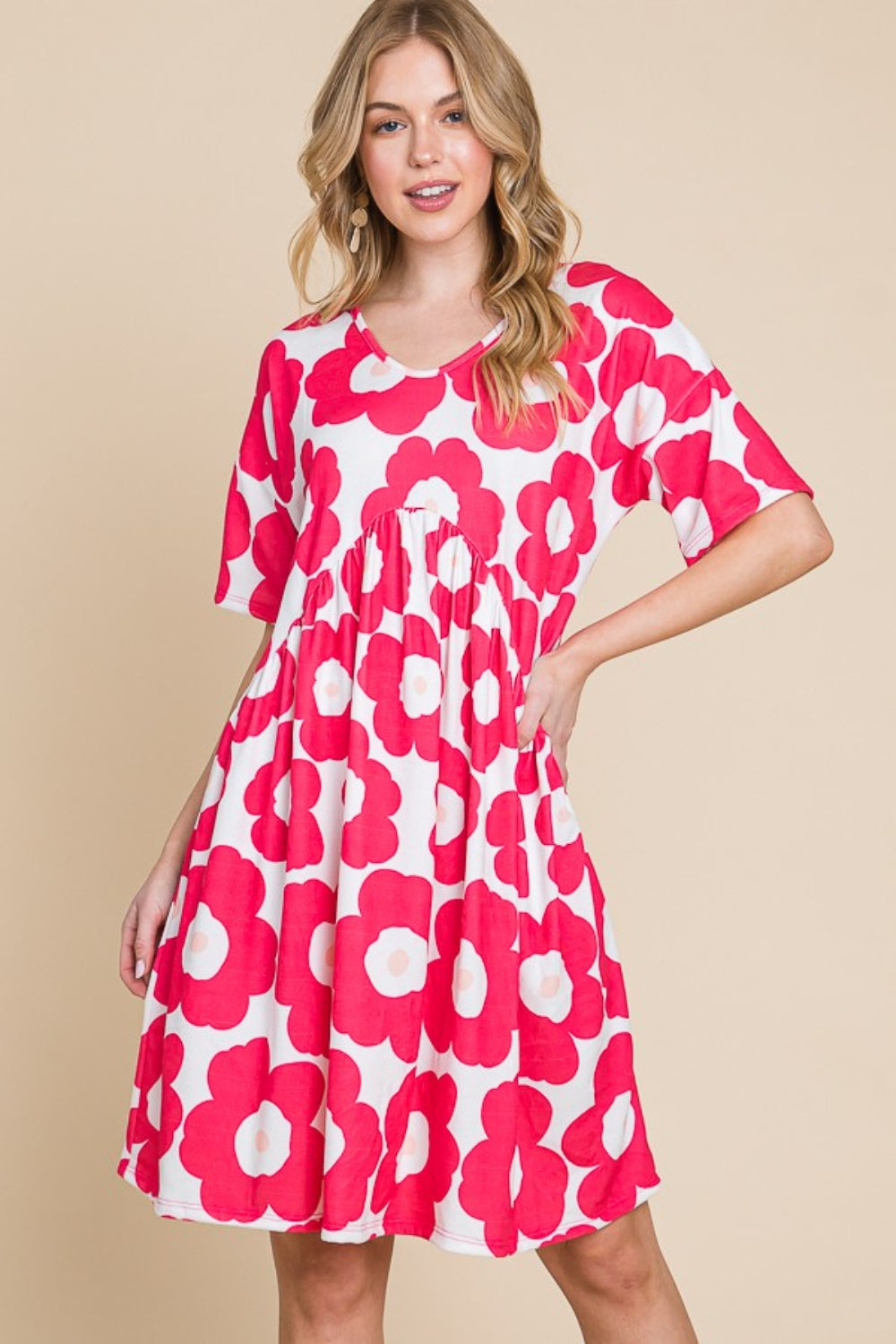 A woman gracefully stands against a beige background, wearing the BOMBOM Flower Print Ruched Dress adorned with large pink blooms. The stretchy fabric of the dress complements her form, effortlessly enhancing its elegant silhouette.