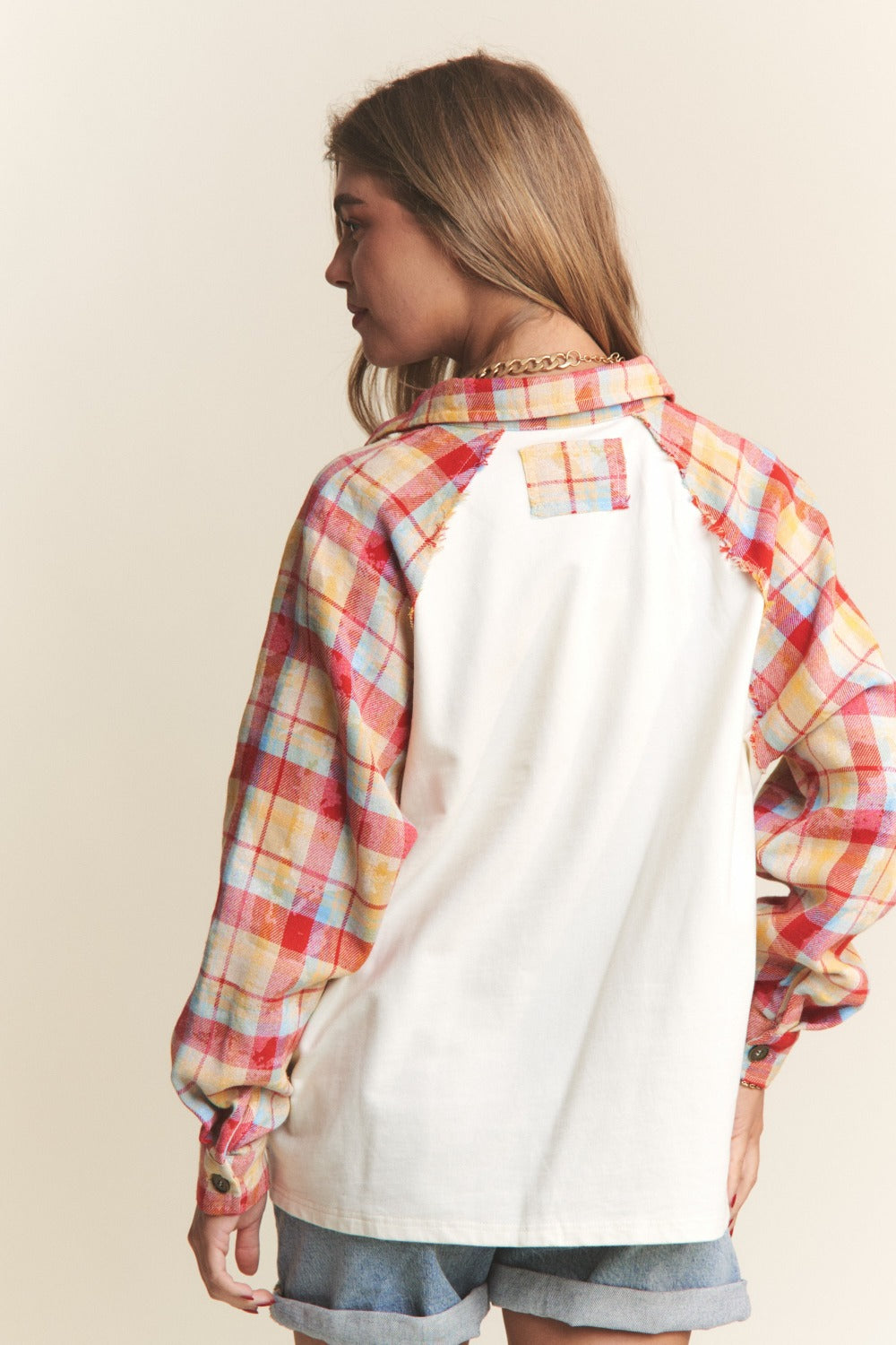 A woman wearing the J.NNA Plaid Contrast Button Down Henley Top—a stylish white pullover shirt with red and yellow plaid sleeves—and denim shorts, stands against a plain background.