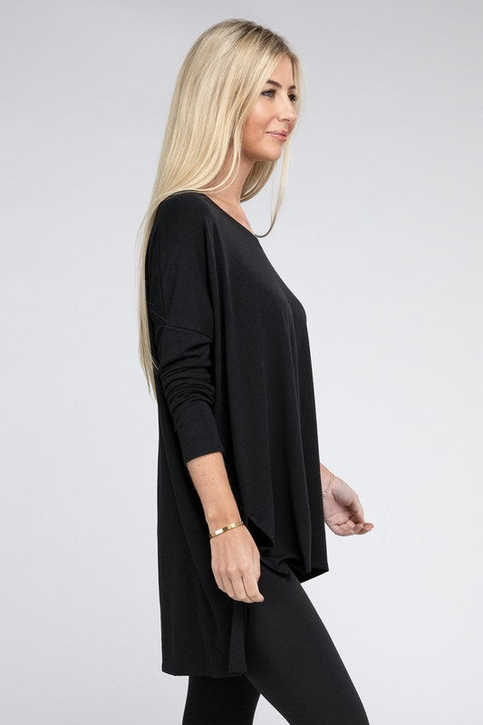 A woman with long, blonde hair is wearing a loose-fitting, brown Dolman Long Sleeve V-Neck Side Slit Hi-Low Hem Top and black pants. She poses with one hand on her hip and a neutral facial expression against a light grey background.