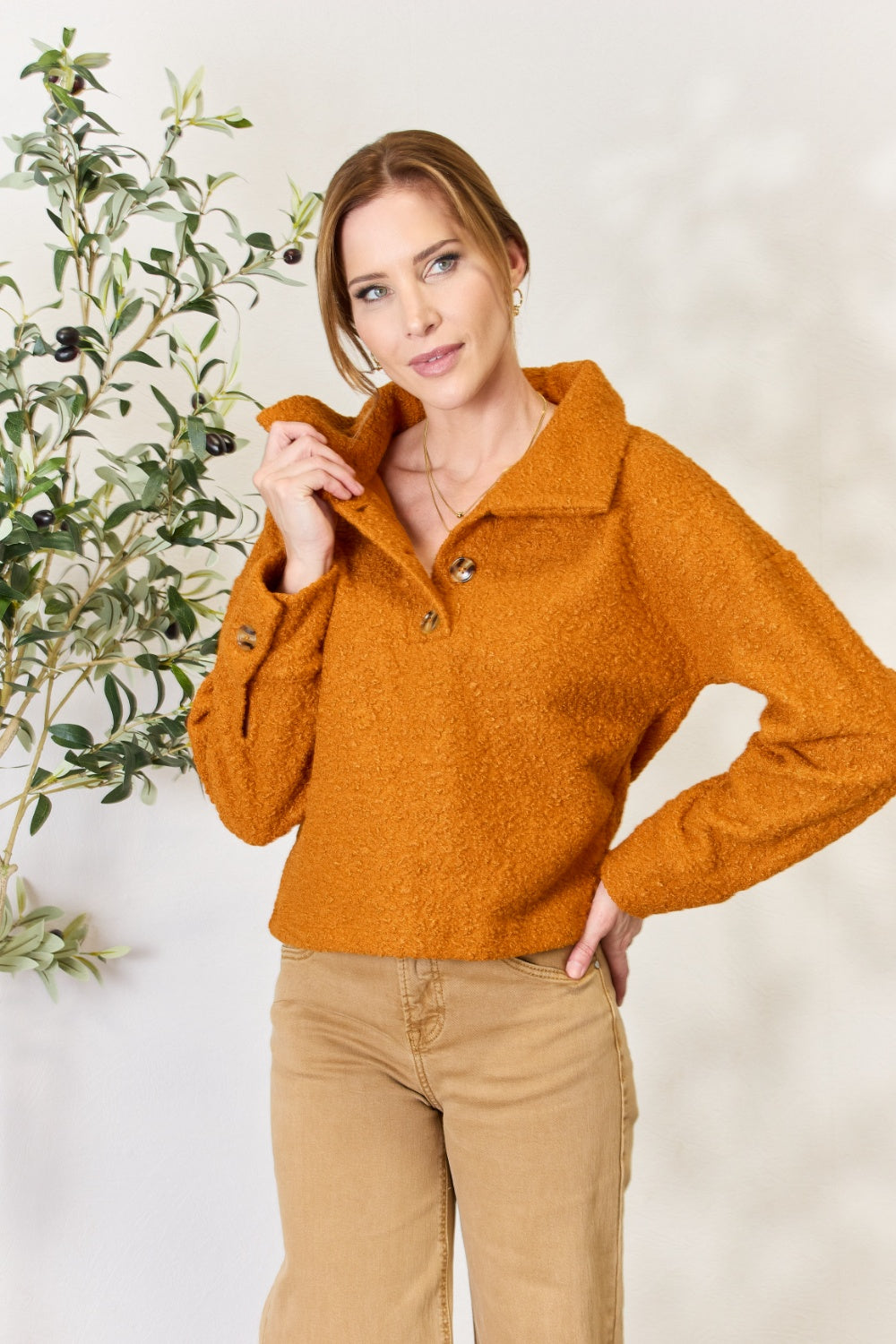 A person wears a brown textured Culture Code Full Size Half Button Turtleneck Sweatshirt made from breathable fabrics and tan pants, standing next to a leafy plant against a light background.