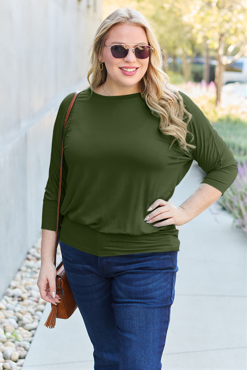 A woman in a slightly stretchy, brown Basic Bae Full Size Round Neck Batwing Sleeve top and blue jeans stands against a concrete wall, holding a brown handbag.