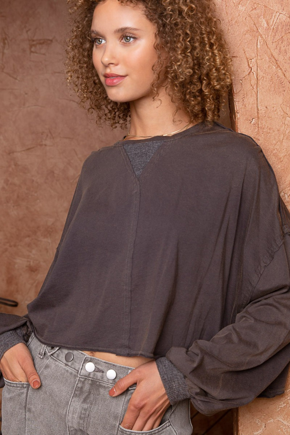 A woman with curly hair is wearing a stylish POL Cropped Long Sleeve Top and gray pants, standing with one hand in her pocket against a textured brown background.