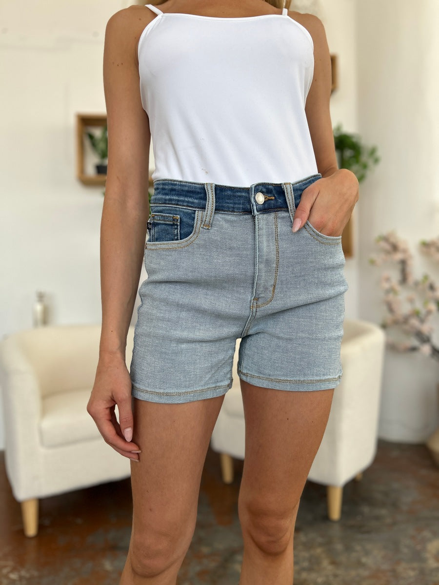 A person models indoors beside a beige chair and a plant, sporting a white t-shirt with the Judy Blue Full Size Color Block Denim Shorts for an effortlessly bold, eye-catching look.