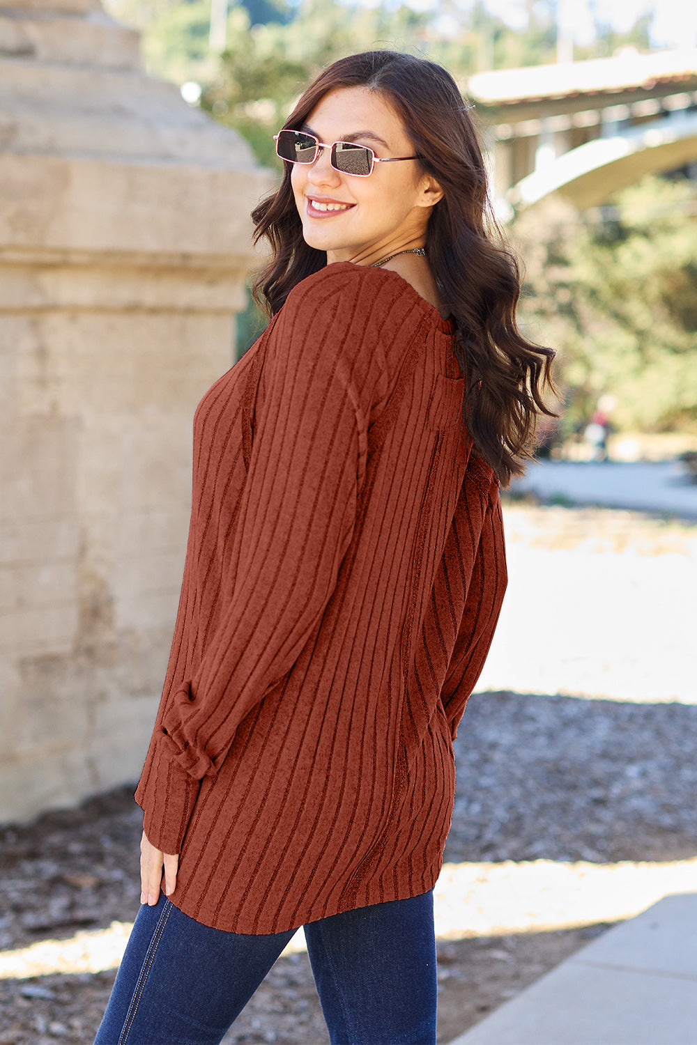 A person with long dark hair and sunglasses stands outdoors, wearing the Basic Bae Full Size Ribbed Round Neck Long Sleeve Knit Top in blue and a pair of jeans. The basic style outfit blends seamlessly with the background of trees and a bridge.