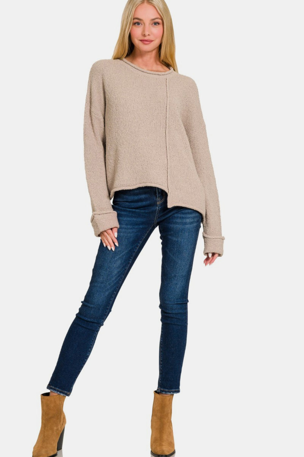 A woman with long blonde hair wears a trendy Zenana Asymmetric Hem Drop Shoulder Sweater in beige and blue jeans, posing with one hand touching her chin and the other holding the hem of her sweater.