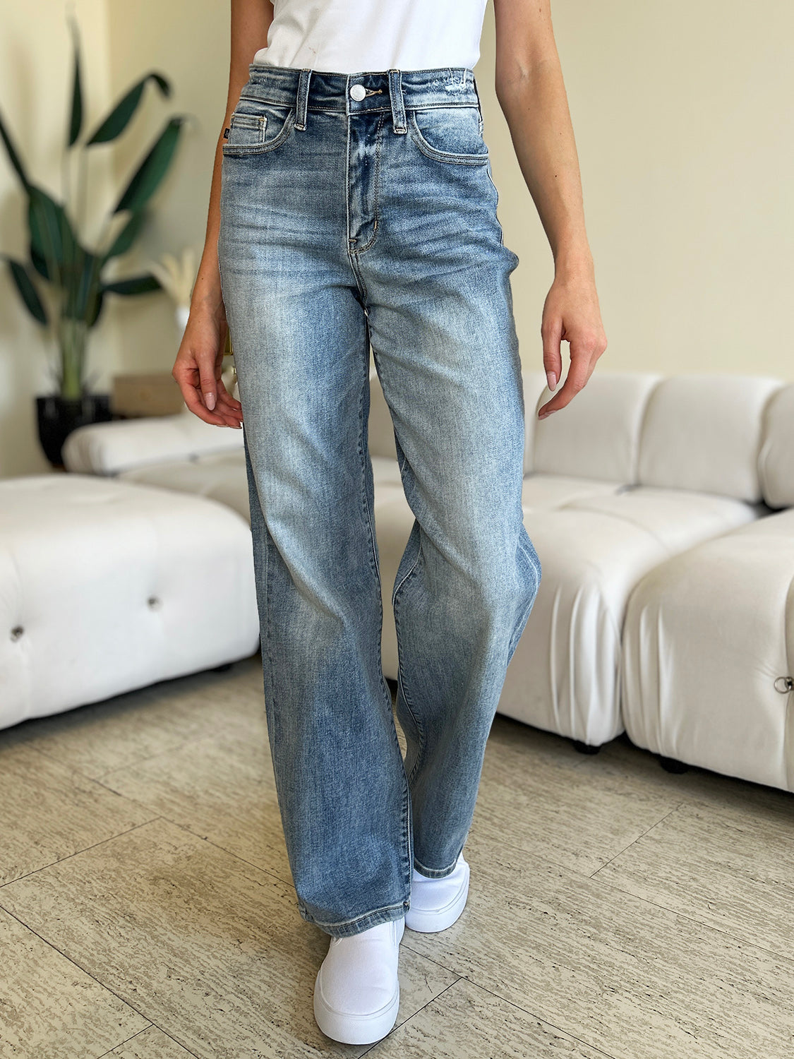 A person wearing Judy Blue Full Size High Waist Straight Jeans and white shoes is standing indoors. There is a white couch and a large plant in the background.