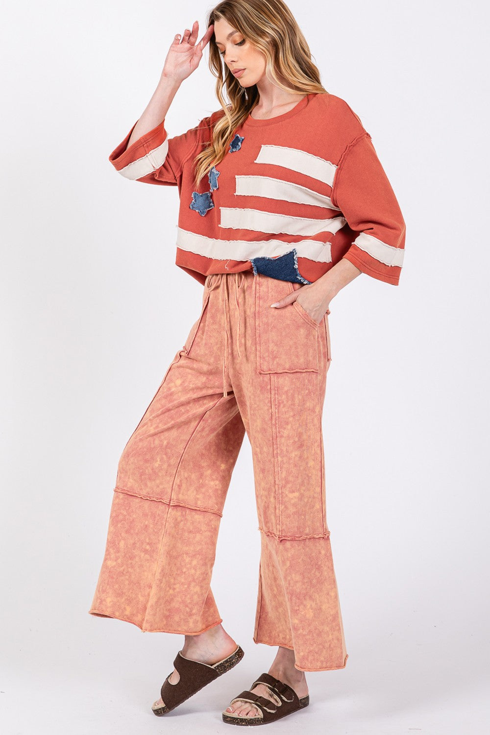 Person wearing SAGE + FIG Mineral Washed Terry Wide Leg Pants with an adjustable waistband and oversized pockets in orange, paired with sandals and a striped top.