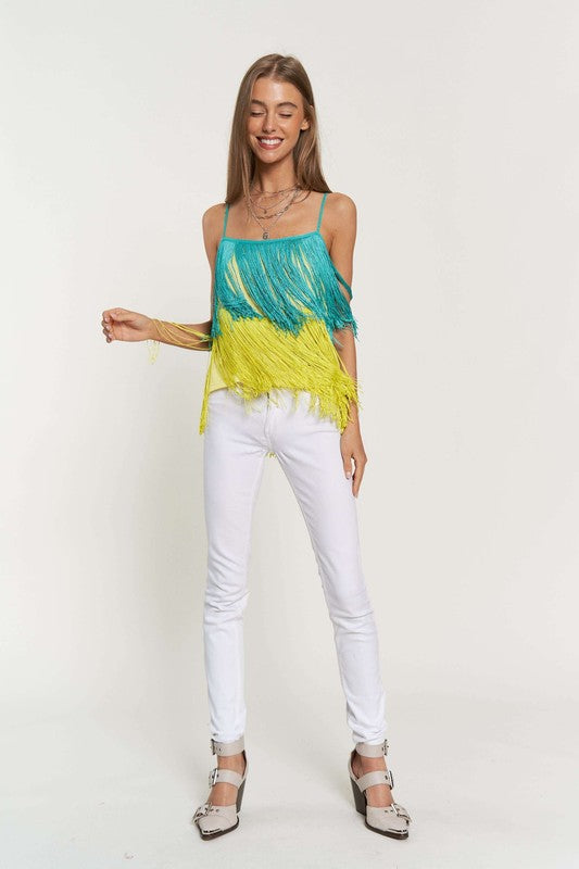 A woman with long brown hair is wearing a Fringe Overlay Cross Straps Party Cami Top featuring blue and yellow layers, paired with white pants, standing against a plain background.