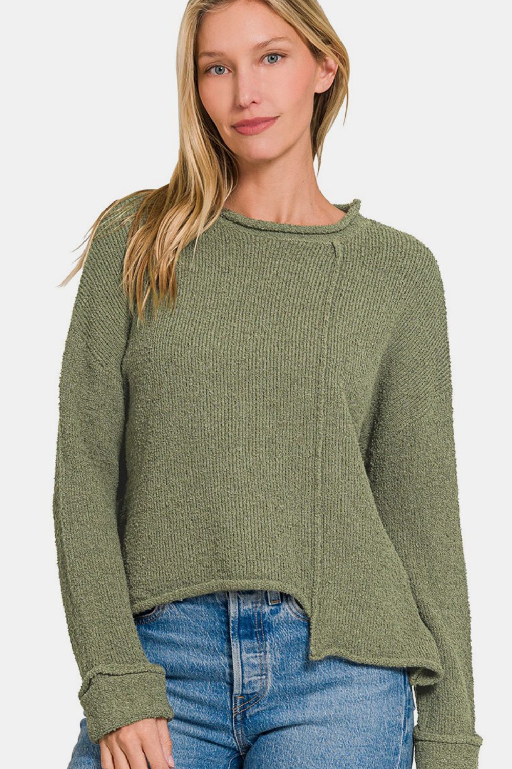 A woman with long blonde hair, dressed in the trendy Zenana Asymmetric Hem Drop Shoulder Sweater in green and blue jeans, poses against a plain background.