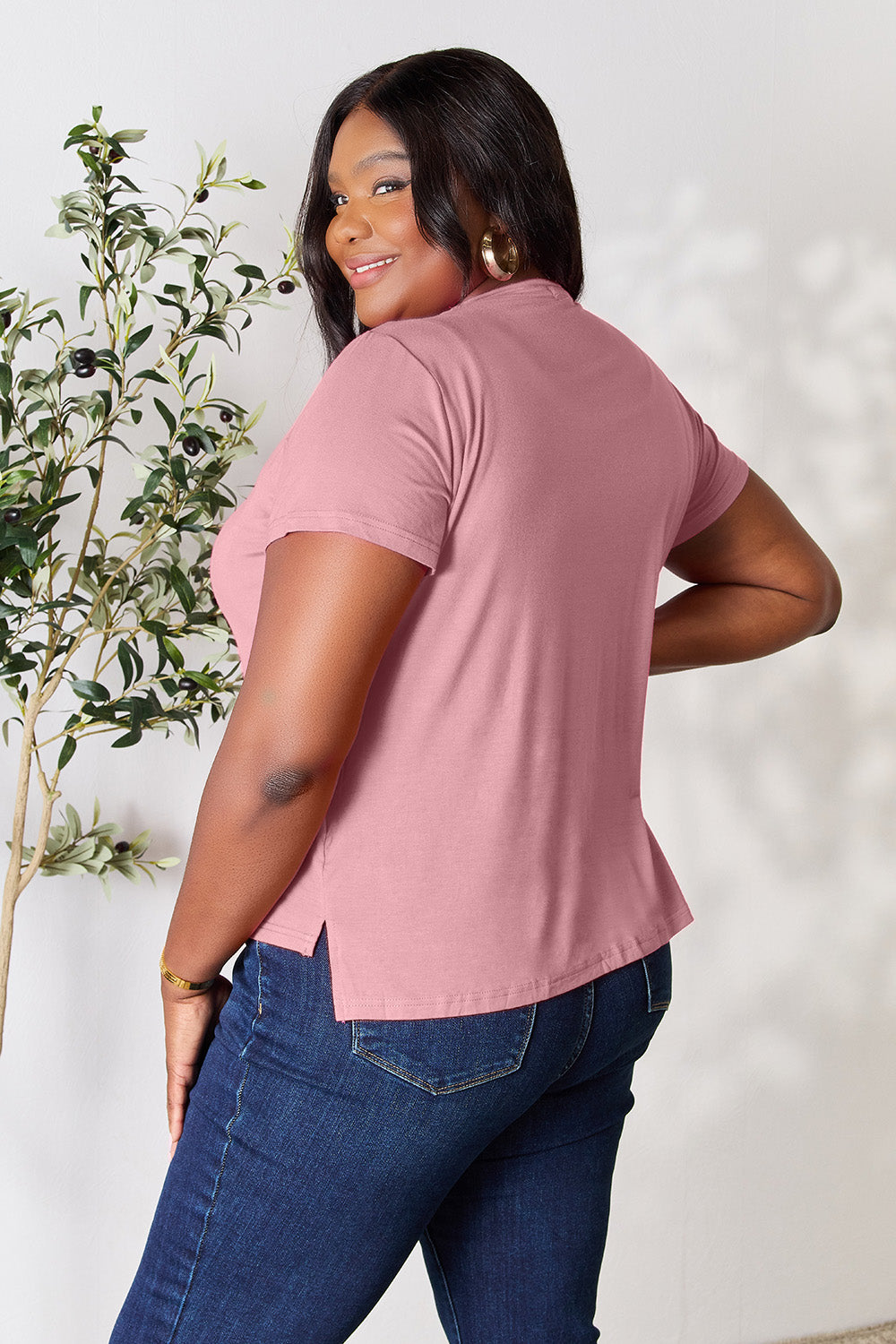 A woman stands next to an indoor plant, smiling, with one hand in her khaki pants pocket. In her pink Basic Bae Full Size Round Neck Short Sleeve T-Shirt and gold jewelry, she radiates casual elegance.