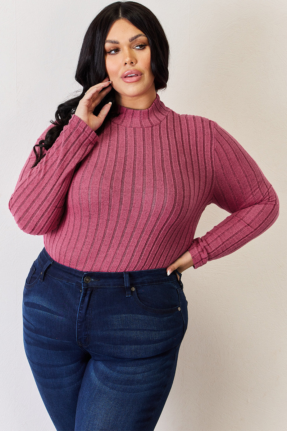 A person with long, wavy blonde hair wears a Basic Bae Full Size Ribbed Mock Neck Long Sleeve T-Shirt in pink stripes and dark jeans made of a comfortable polyester blend, standing in front of a white curtain.