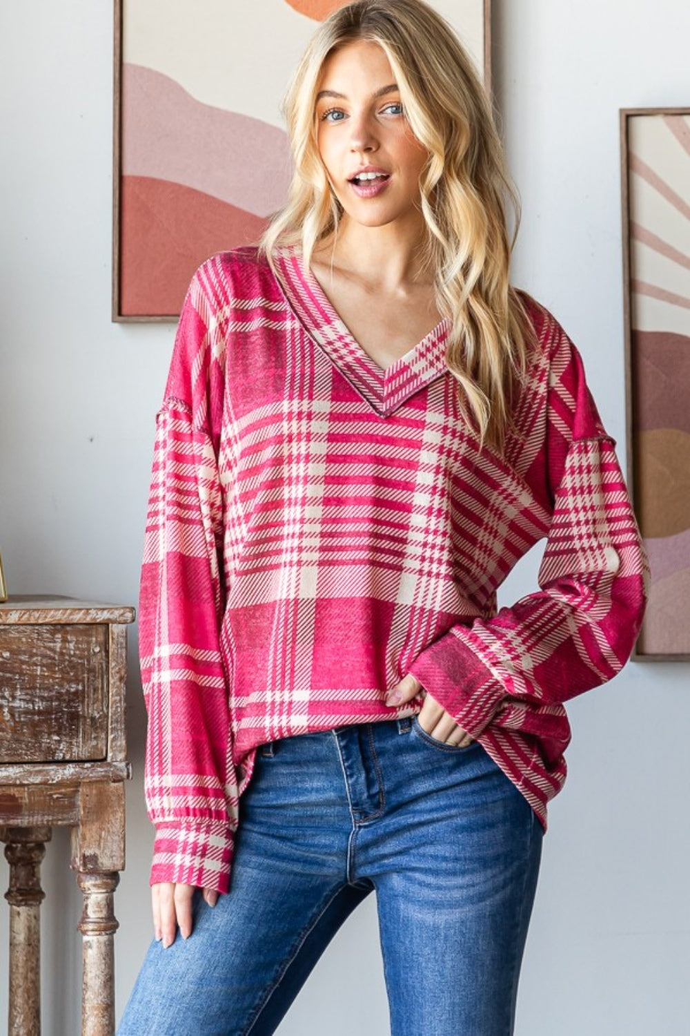 A person with long blond hair smiles while wearing the Heimish Plaid Drop Shoulder V-Neck T-Shirt in red and white and a pair of blue jeans, leaning against a wooden table with framed pictures in the background.