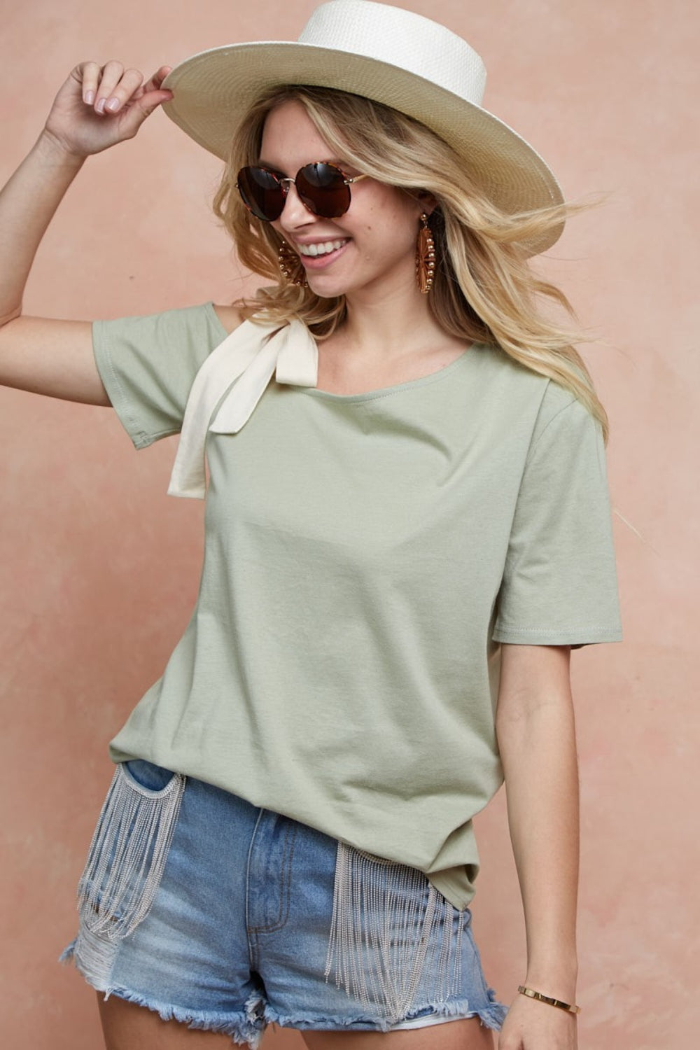 A person smiling, sporting a wide-brimmed hat, sunglasses, and the BiBi Tied Ribbon One Shoulder Short Sleeve T-Shirt in light green, paired with denim shorts, stands against a peach background.