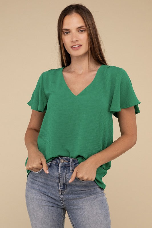 A person stands against a beige background, wearing a green Woven Airflow Flutter Sleeve Top with chic breathable fabric and high-waisted blue jeans. Hands are in their pockets, and they have long brown hair.