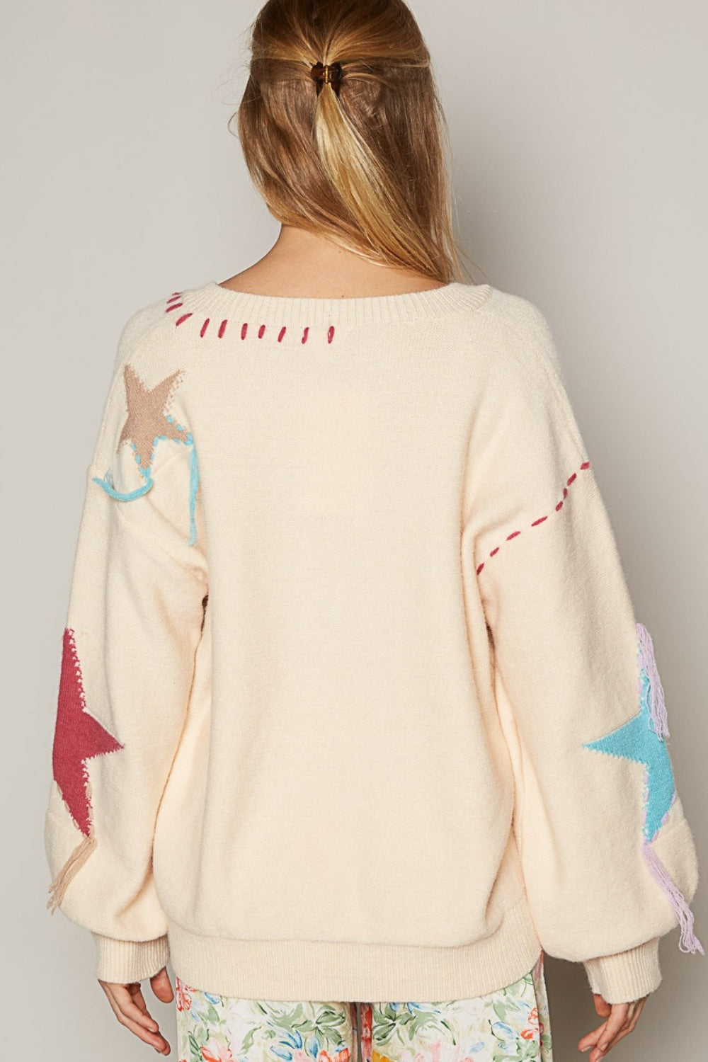 Person wearing a cozy POL V-Neck Long Sleeve Star Fringe Sweater and floral pants, sitting against a plain background.