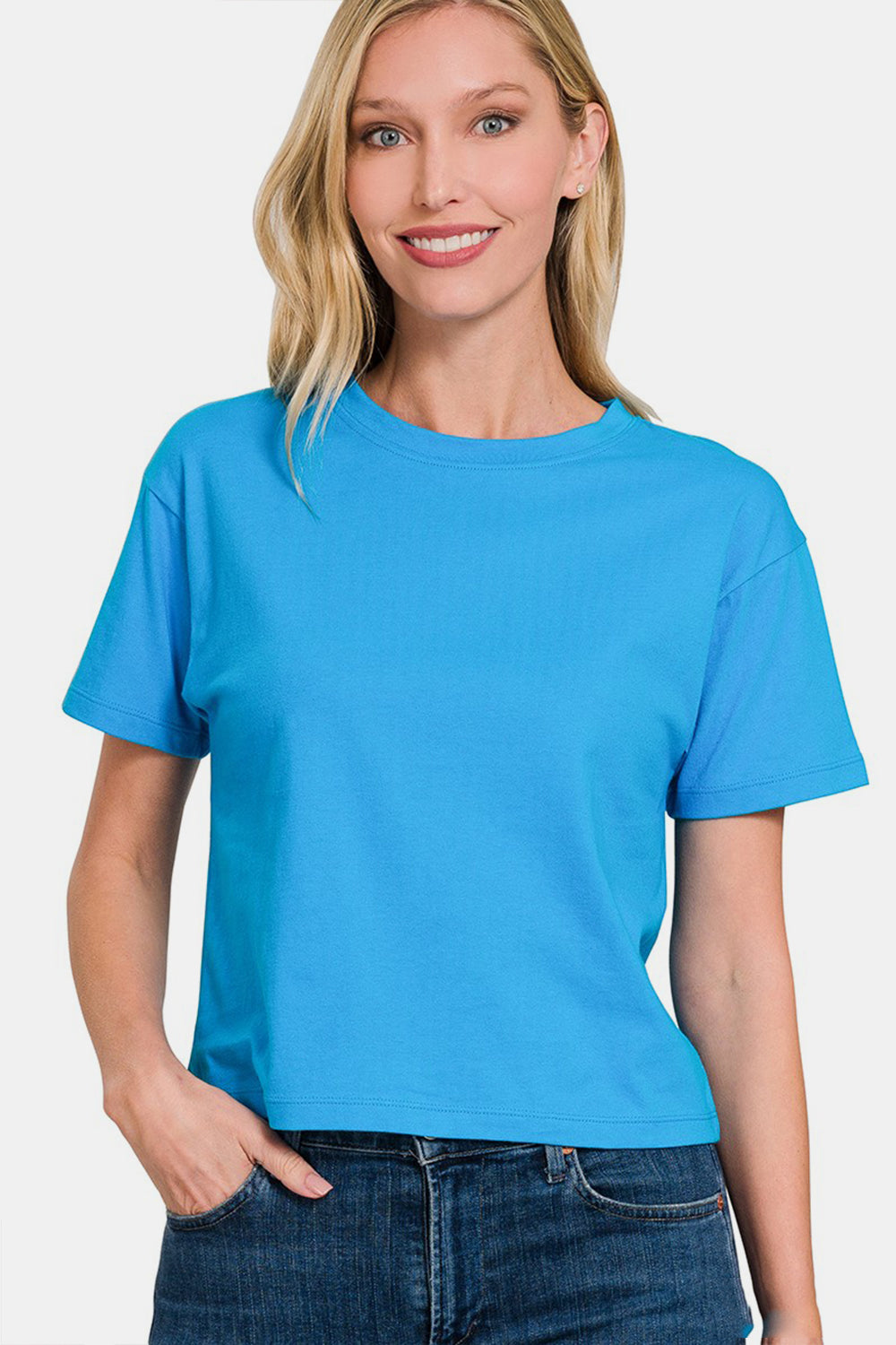 A person with long blonde hair is smiling while wearing the Zenana Round Neck Short Sleeve Cropped T-Shirt in blue and high-waisted jeans, standing against a plain background.