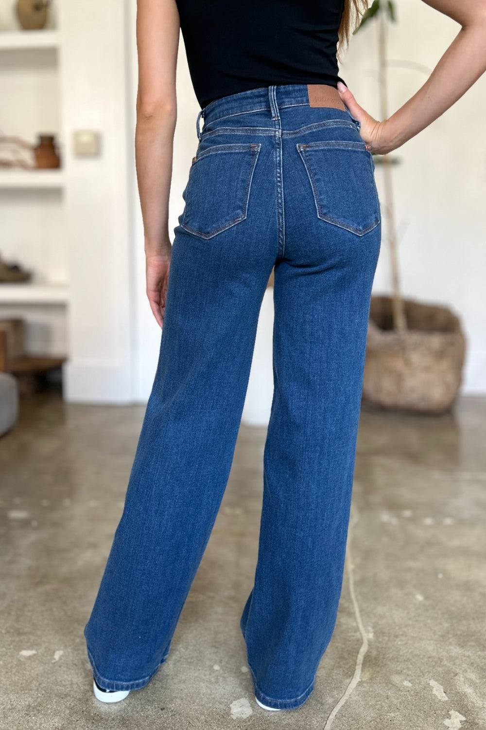 A person wearing Judy Blue Full Size High Rise Straight Jeans, a black top, and white shoes stands gracefully on a polished concrete floor.