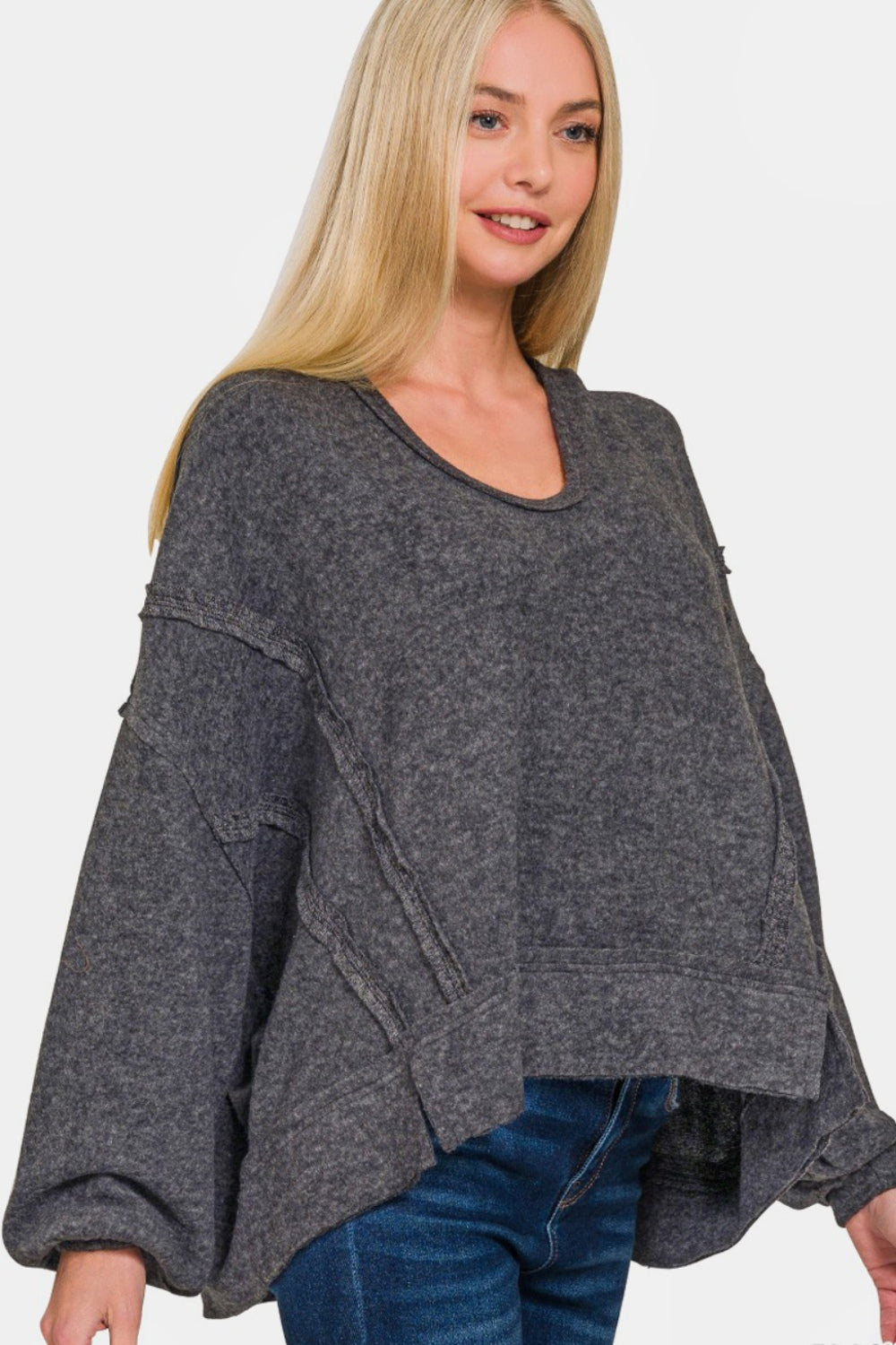 A person with long blond hair stands against a white background, wearing the Zenana Brushed Hacci Exposed Seam Hoodie in dark gray paired with jeans.