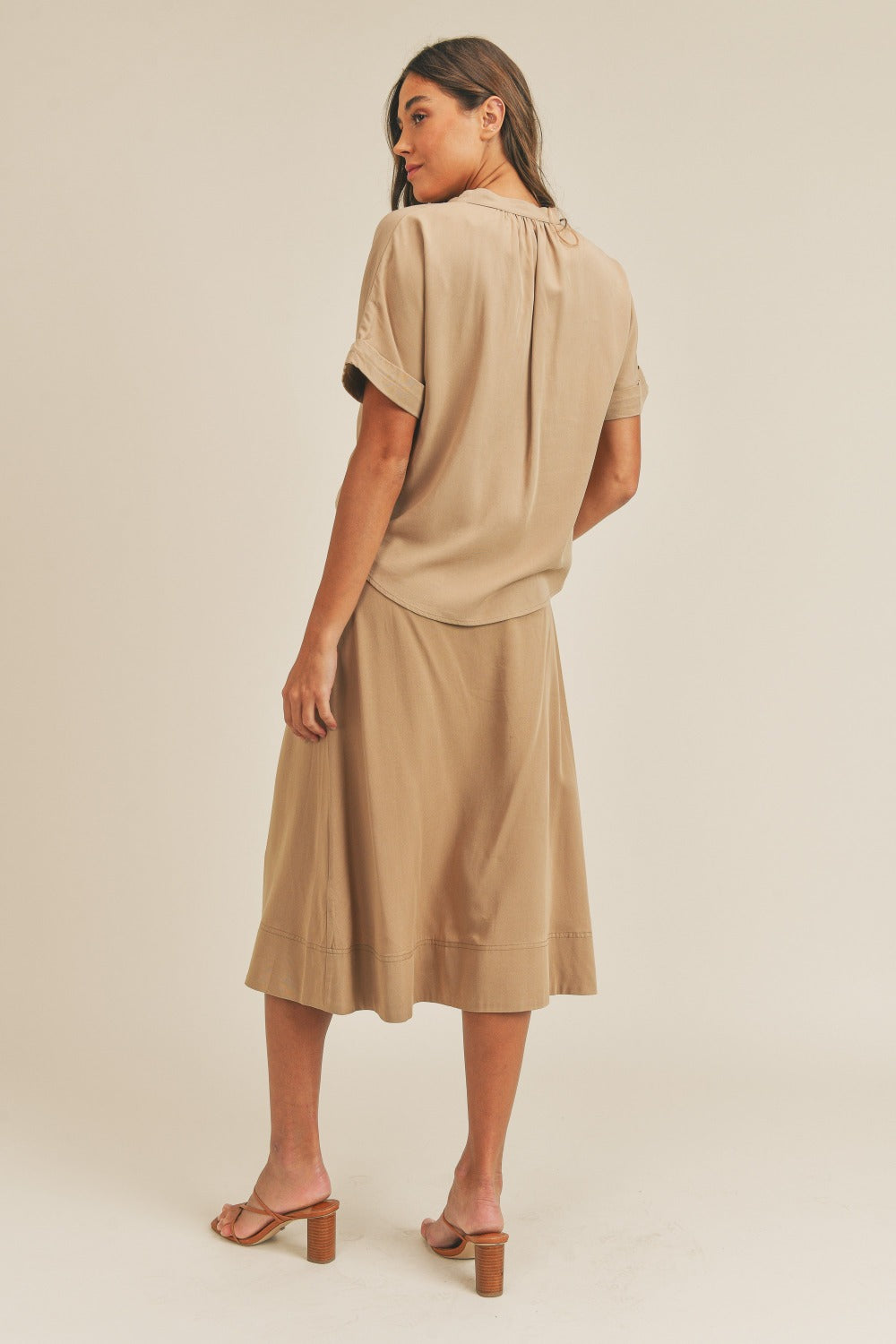 A woman is standing against a plain background wearing the MABLE Short Sleeve Top and Button Down Midi Skirt Set in beige. The elegant two-piece set, which includes a belted midi skirt, is paired perfectly with beige heels as she confidently poses with one hand on her hip.