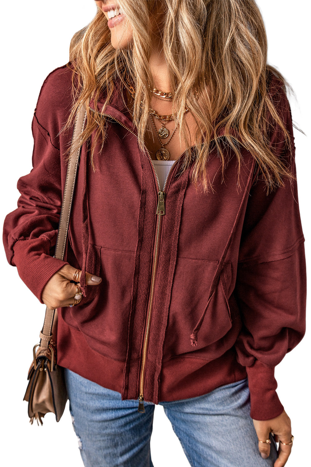 A woman with long, wavy blonde hair is seen from behind, wearing a trendy Brown Raw Edge Exposed Seam Full Zip Hoodie and carrying a brown shoulder bag.