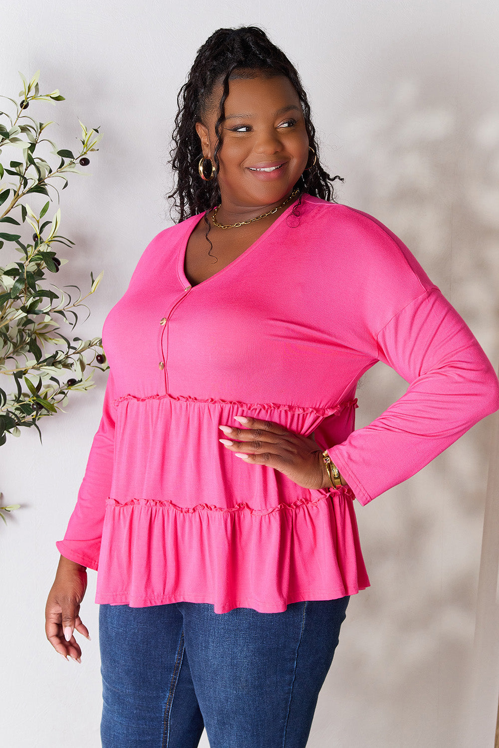 Person stands indoors near a plant, wearing the Double Take Half Button Long Sleeve Ruffle Hem Blouse in gray and blue jeans, smiling while looking to the side. The slightly stretchy fabric of the blouse adds comfort while its buttoned design offers a touch of elegance.