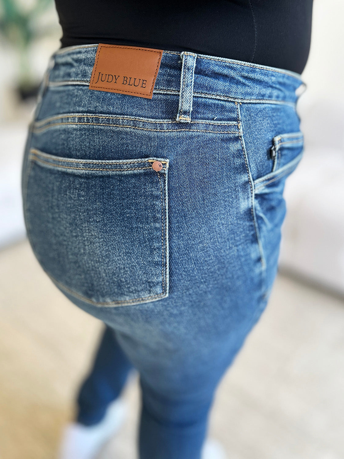 A person wearing Judy Blue Full Size High Waist Skinny Jeans made of stretchy denim fabric stands in a room with light-colored furniture and wooden flooring, paired perfectly with white slip-on shoes.