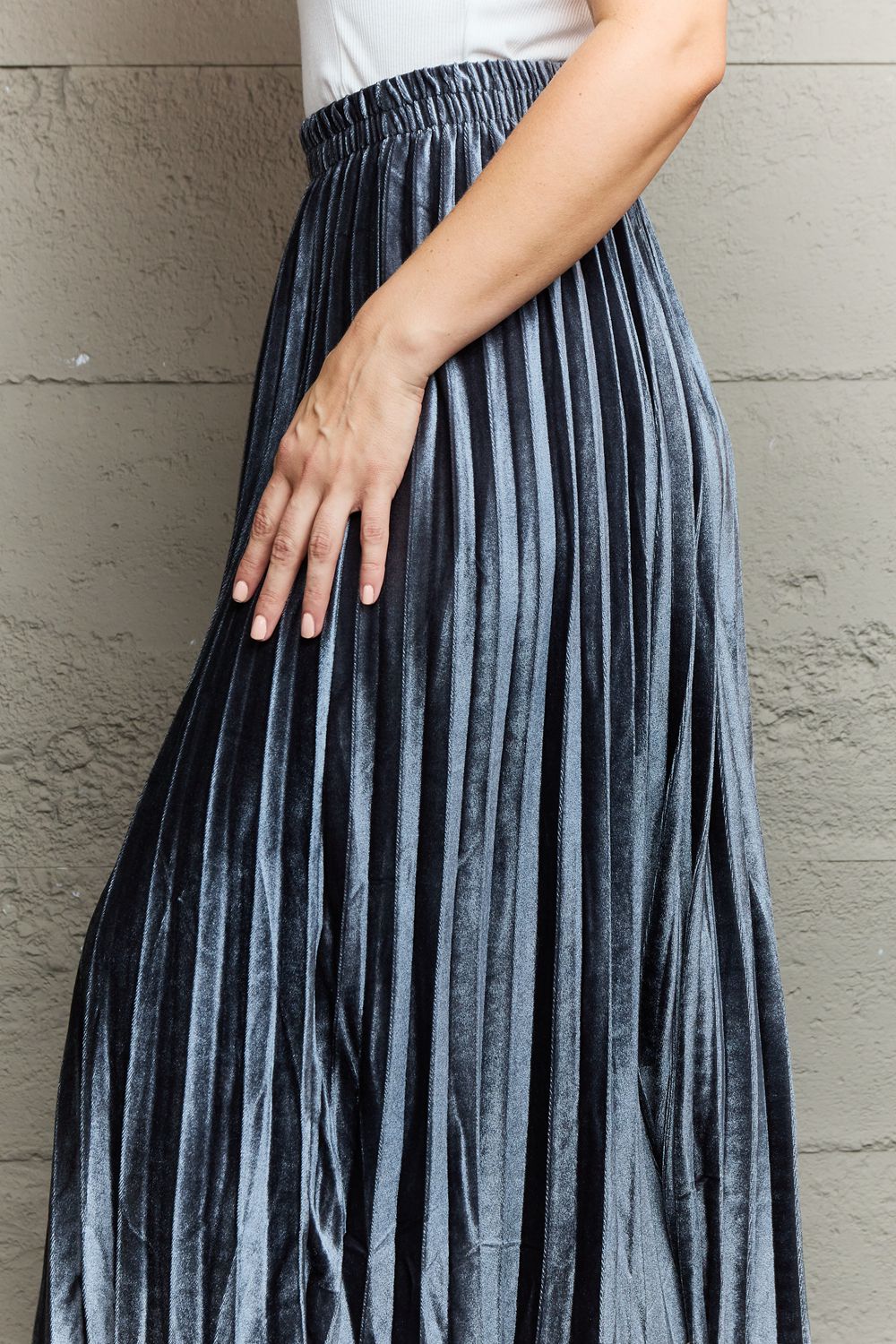 A person in a white top and the Ninexis Accordion Pleated Flowy Midi Skirt stands against a textured wall, wearing beige heeled sandals.