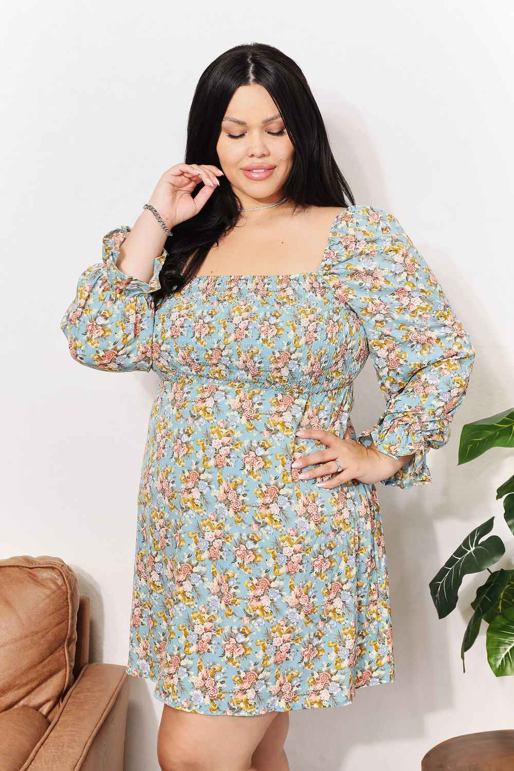 A woman in the Honey Floral Smocked Flounce Sleeve Square Neck Dress stands indoors, holding the hem of her dress. She is smiling slightly with her eyes looking downward. Nearby, a plant and a brown couch complete the cozy scene.