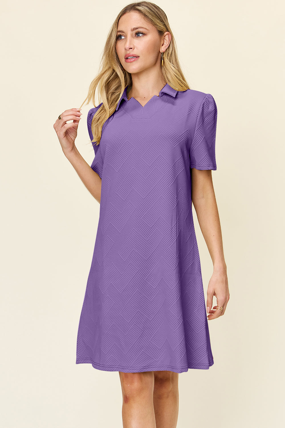 A woman wearing the Double Take Full Size Texture Collared Neck Short Sleeve Dress, featuring a geometric pattern in purple and made from soft polyester, stands against a neutral background. She has long, light hair and is looking slightly to the side. The dress is available in sizes S-3XL and is easy to care for—just machine wash cold.