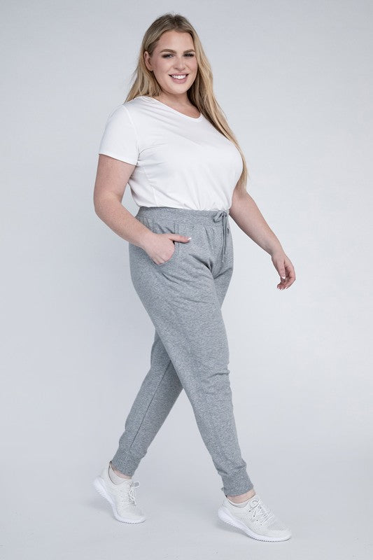 A person stands against a white background, wearing a white t-shirt, grey Plus-Size Jogger Pants with an adjustable waistband, and white sneakers. They have long blonde hair and are posing with one hand on their hip.