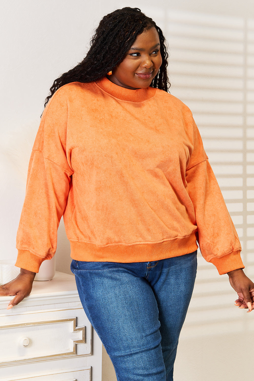 A woman with long, wavy hair smiles in a casual wardrobe, sporting the Basic Bae Round Neck Dropped Shoulder Sweatshirt in orange and blue jeans. She stands beside a white dresser adorned with decorative vases.