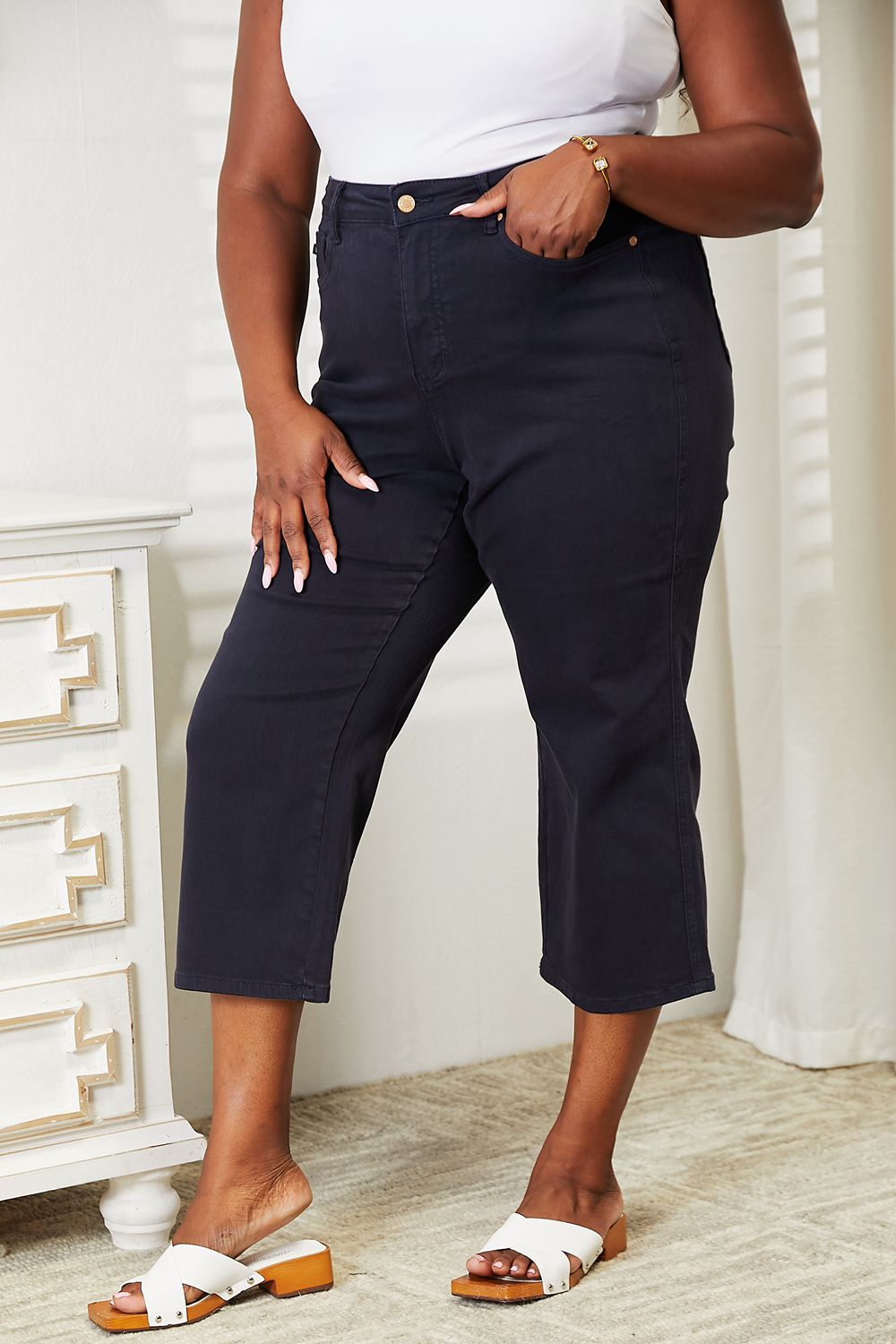 A person wearing a white sleeveless top and Judy Blue Full Size High Waist Tummy Control garment-dyed wide cropped jeans in black stands indoors next to a white dresser.