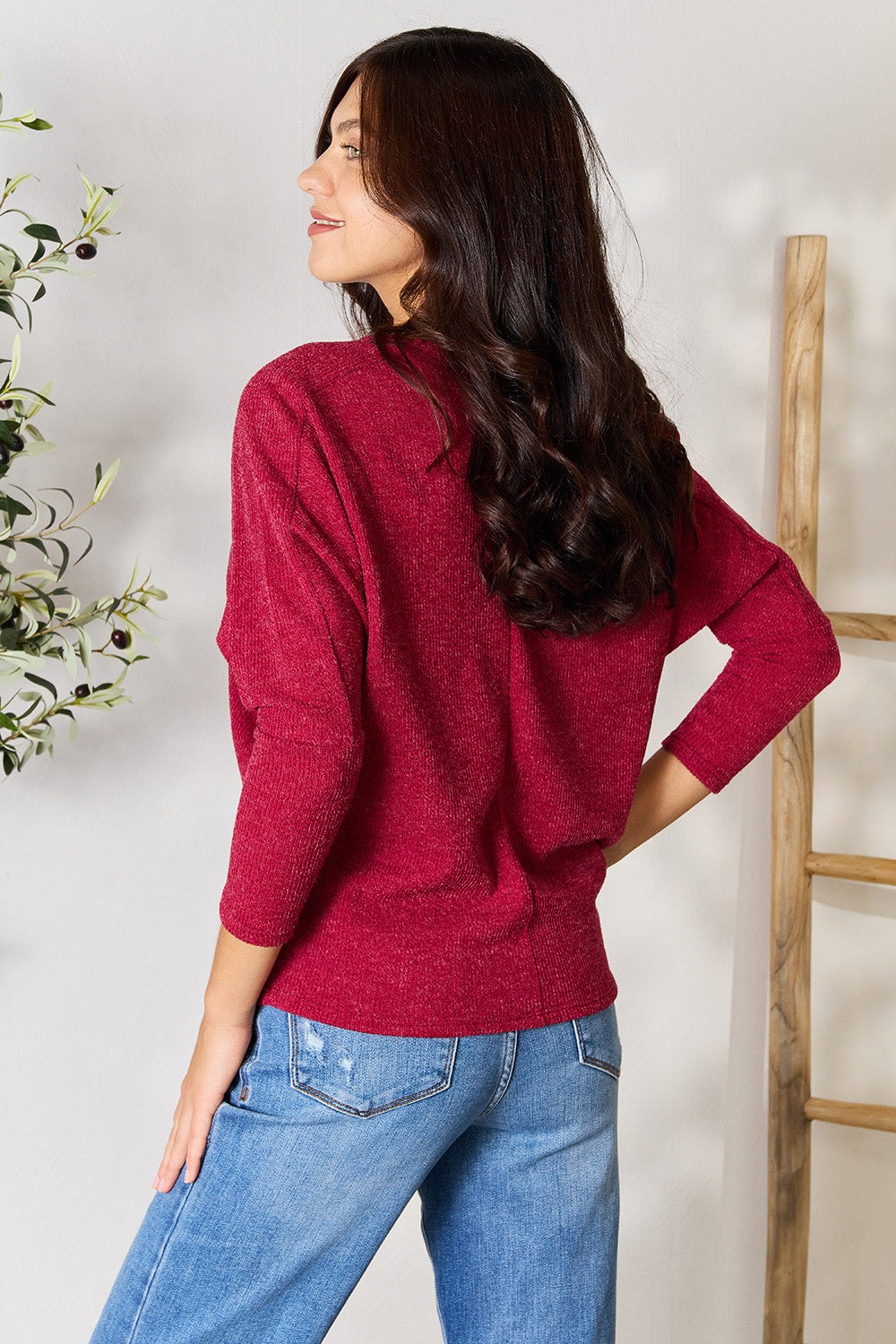 Person wearing a BOMBOM Drop Shoulder Long Sleeve Blouse in red, paired with blue jeans, standing with one hand in their pocket and the other touching their neck, in front of a white wall with a potted plant on the side.