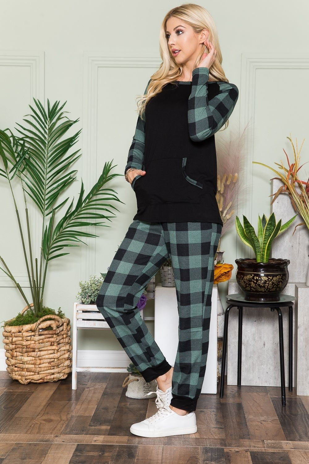 An individual wearing a Celeste Plaid Long Sleeve T-Shirt with Pockets stands indoors on a wooden floor, next to potted plants and a wicker basket.