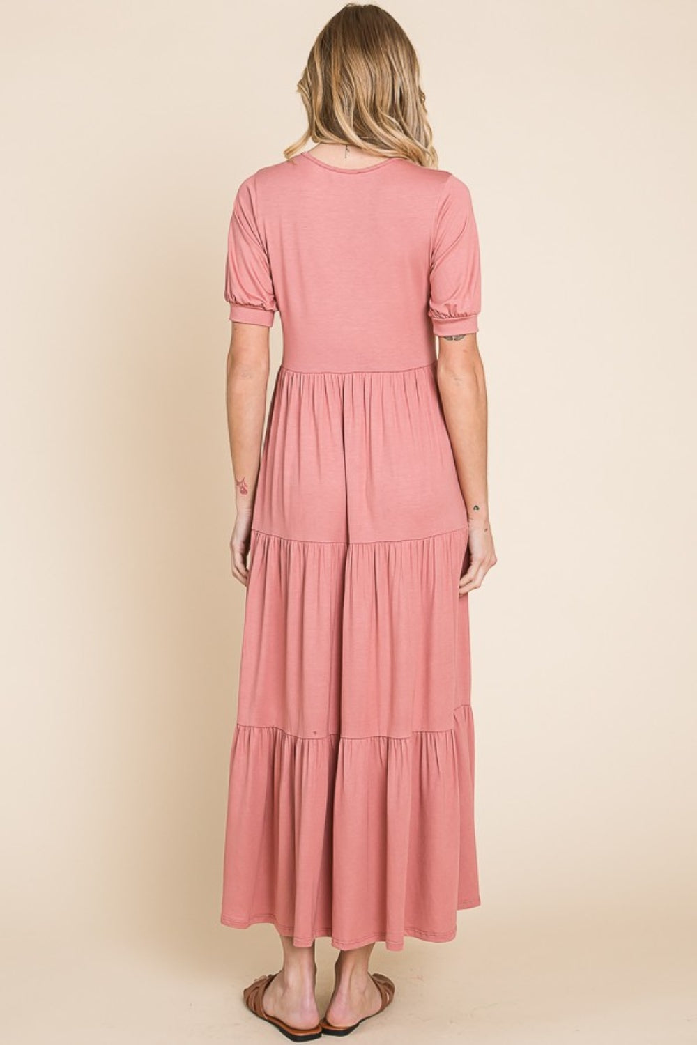 A person wearing the BOMBOM Short Sleeve Tiered Maxi Dress in pink, showcasing its comfortable style with a long, tiered design, stands against a beige background.