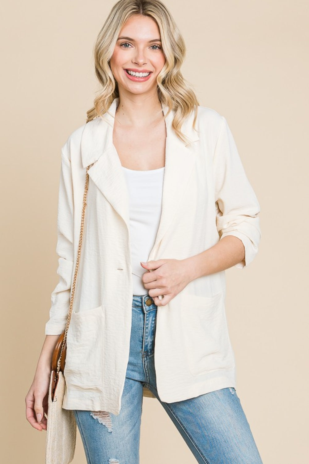 A person with long hair wearing the Culture Code One Button Long Sleeve Blazer with Pockets in light cream stands against a beige background, smiling. The blazer's tailored fit perfectly complements the white top and blue jeans, creating a versatile look for any occasion.