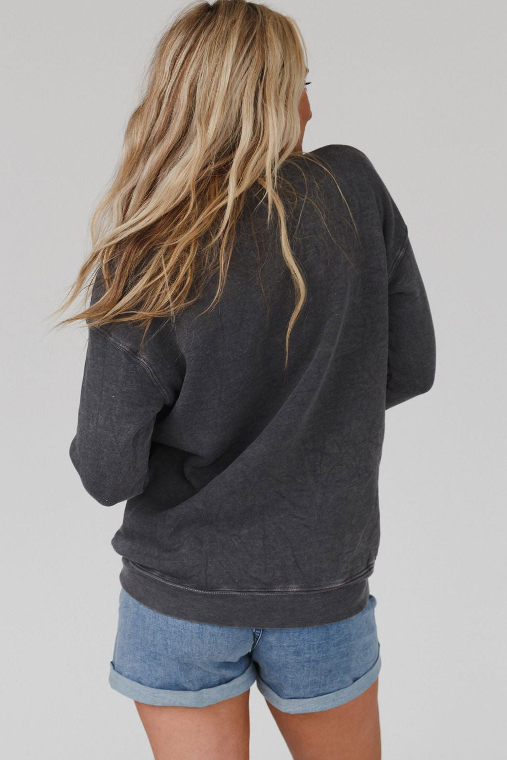 A person with long blond hair wearing a gray COOL MOMS CLUB drop shoulder sweatshirt and relaxed-fit blue jeans is seen from the back, standing near a pumpkin and a window.