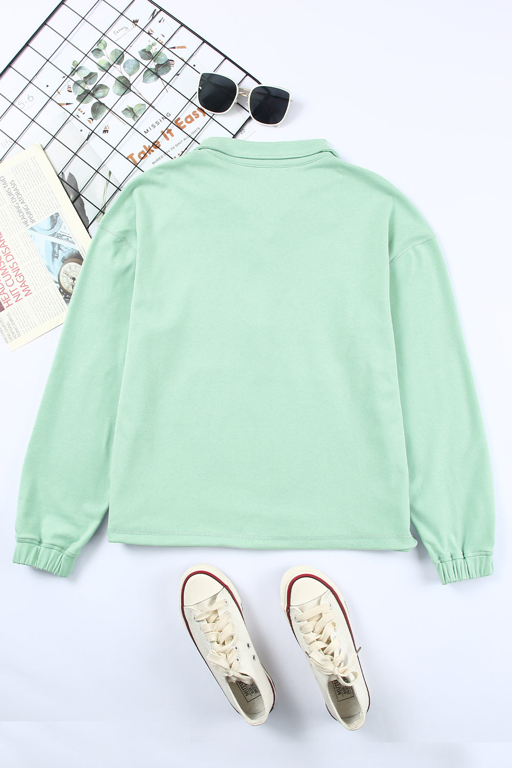 A person with wavy hair stands with their back to the camera, wearing a Green Zip Front Pocketed Pullover Sweatshirt and blue jeans, holding a small brown bag in their left hand.