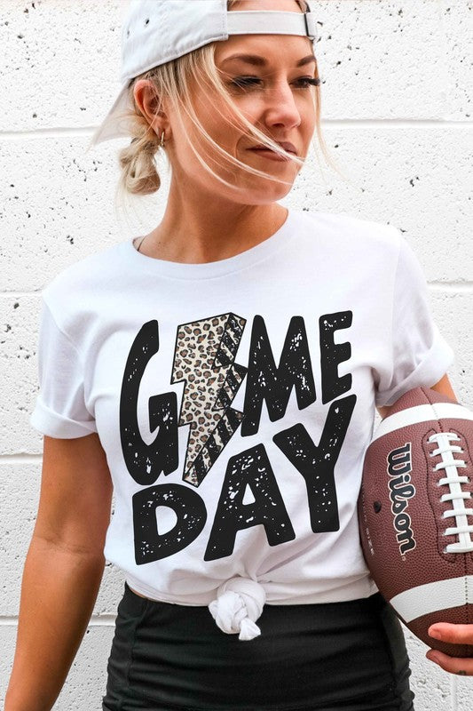 A person wearing a white "LEOPARD LIGHTNING GAME DAY Graphic Tee" with a leopard print lightning bolt design stands with a denim jacket partially on, showcasing its unisex sizing.