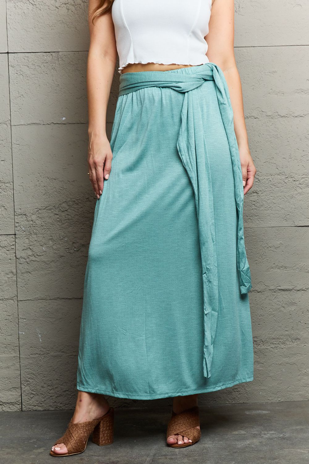 A woman stands against a textured gray wall, wearing the Ninexis Know Your Worth Criss Cross Halter Neck Maxi Dress in teal, featuring a midriff cutout and criss-cross pattern. She pairs it with tan heeled sandals and poses with her right hand on her hip.