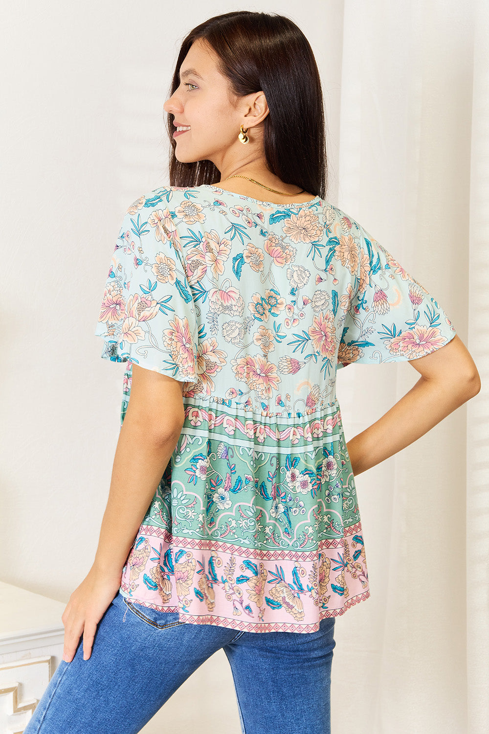 A person wearing a Perfee Floral Tie Neck Short Sleeve Blouse and denim jeans stands indoors, smiling slightly.