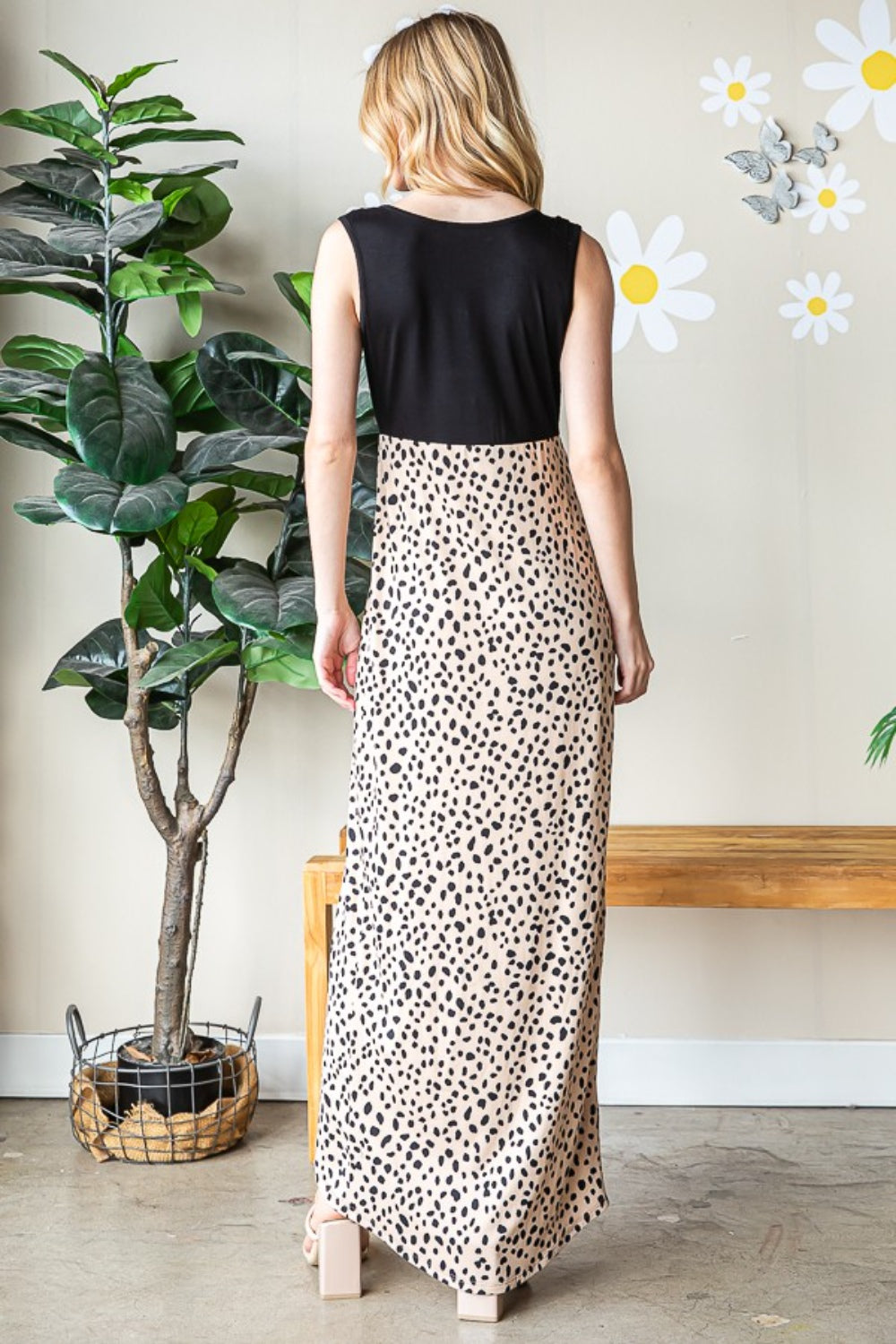 A woman poses indoors wearing the Heimish Full Size Slit Animal Print V-Neck Wide Strap Dress. With one hand resting on her head, she stands elegantly surrounded by potted plants and wall decorations.