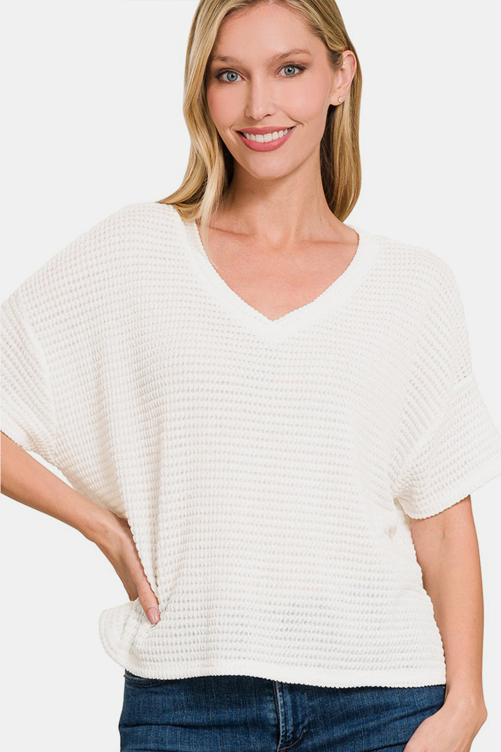 A woman wearing a Zenana Drop Shoulder Short Sleeve Jacquard Knit Top in white stands with one hand on her hip and smiles. She has long, light brown hair and is paired with dark blue jeans.