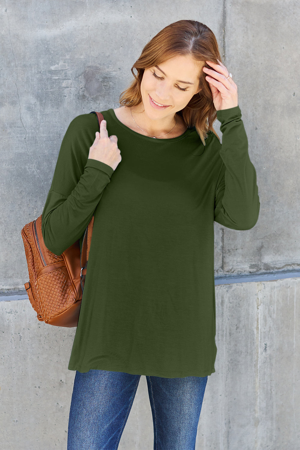 A woman with shoulder-length hair wearing a Basic Bae Full Size Round Neck Dropped Shoulder T-Shirt in green and a pair of jeans, exuding a basic style, stands against a concrete wall. She's holding an imported brown woven backpack on her shoulder while touching her hair with her hand.