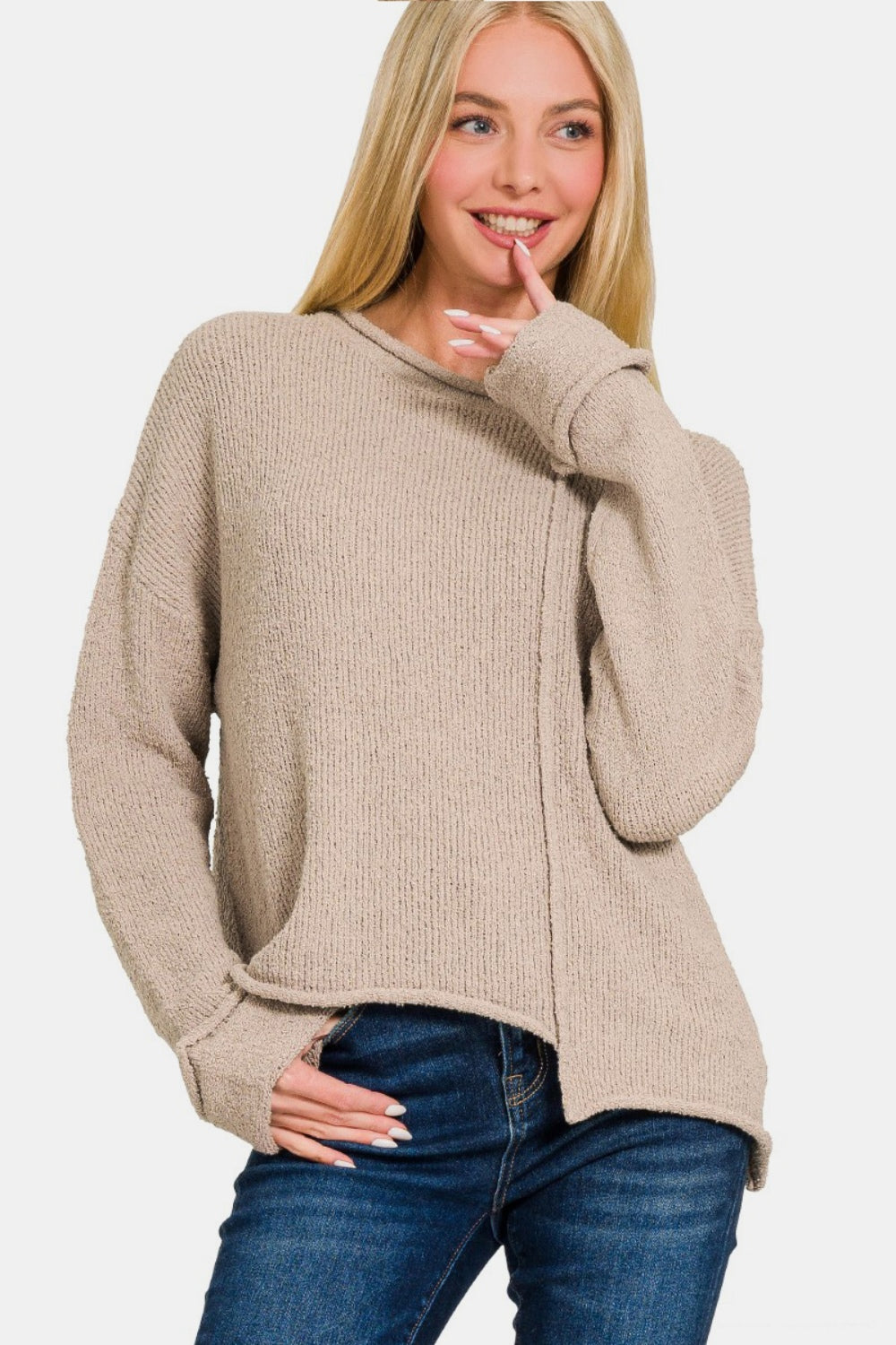 A woman with long blonde hair wears a trendy Zenana Asymmetric Hem Drop Shoulder Sweater in beige and blue jeans, posing with one hand touching her chin and the other holding the hem of her sweater.