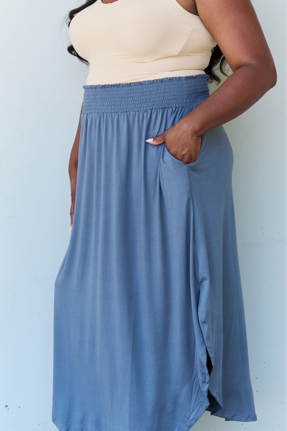 A person wearing a white sleeveless top and the Doublju Comfort Princess Full Size High Waist Scoop Hem Maxi Skirt in Charcoal stands against a plain wall. They are also wearing black sandals.