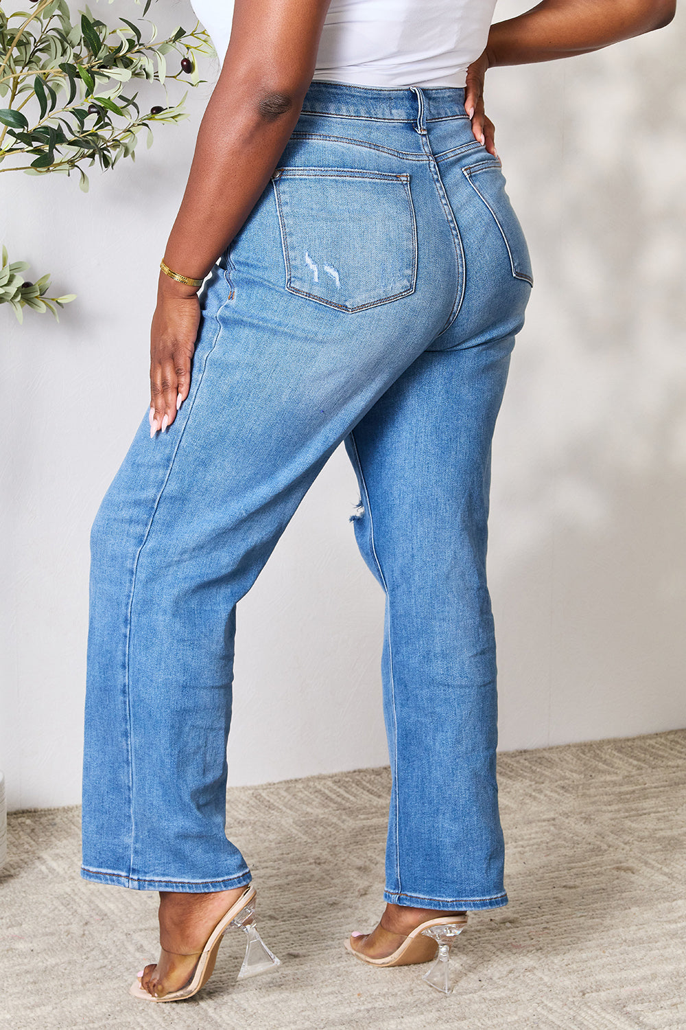 A person wearing a white top and Judy Blue Full Size High Waist Distressed Jeans stands in a room with a ladder and a plant.