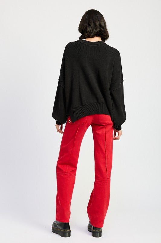 A woman wearing an OVER FITTED LONG SLEEVE SWEATER TOP made of 60% cotton in brown and khaki shorts holds a denim handbag with a brown strap against a neutral background.