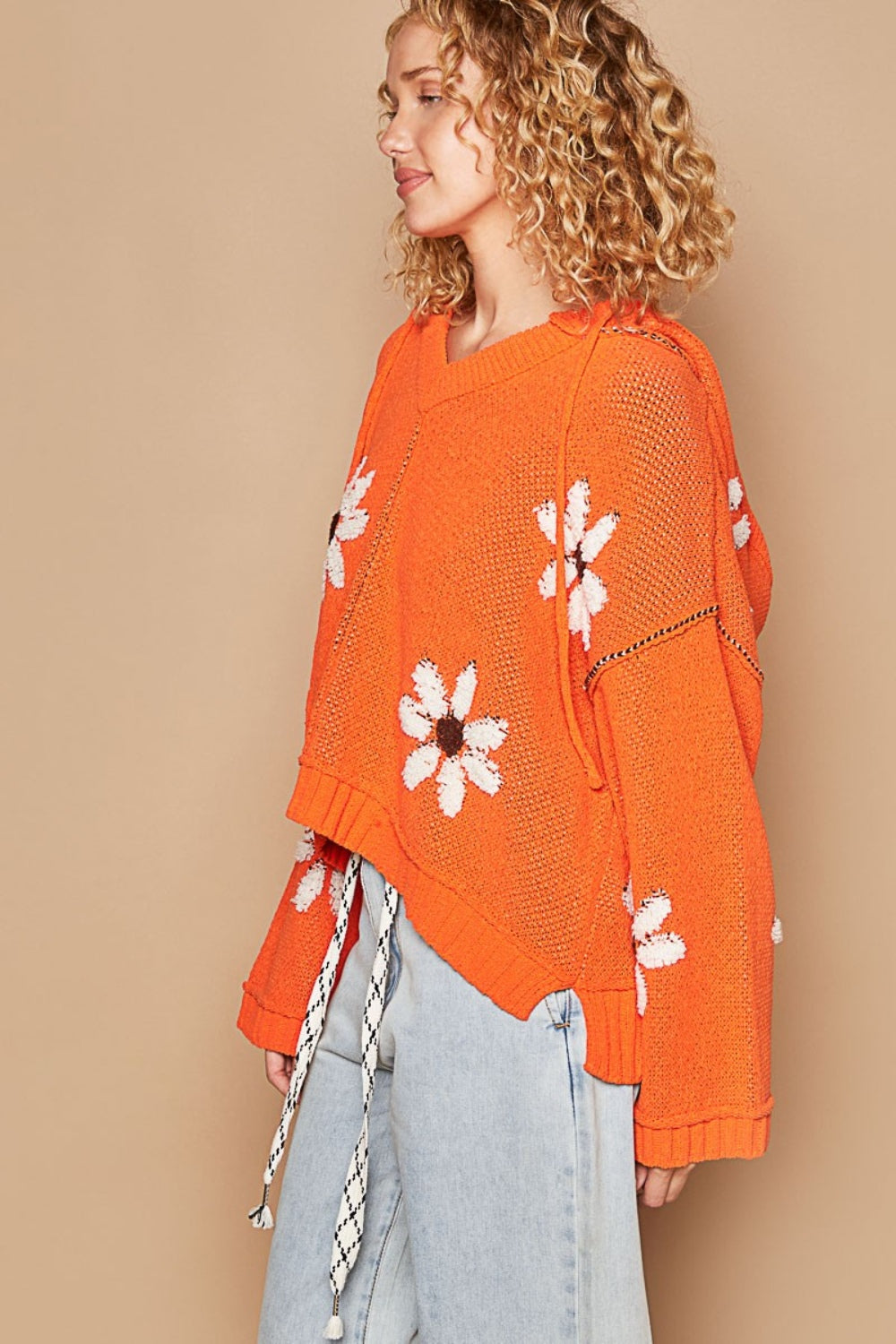 A person with curly blond hair is wearing the POL Floral Pattern Hooded High-Low Sweater in orange, featuring white and brown flower patterns against a solid beige background, paired with light blue jeans.